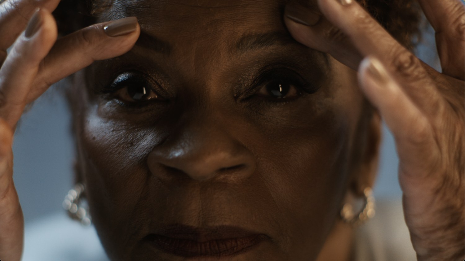 A close up of an older woman, hands at her temples